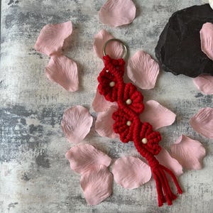 May include: Red macrame keychain with a tassel and wooden beads, shaped like flowers. The keychain is photographed against a white background with pink flower petals.