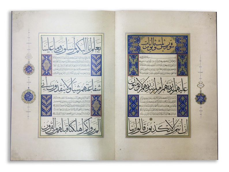 K&ouml;nnte beinhalten: Ein aufgeschlagenes Buch mit zwei Seiten komplexer arabischer Kalligrafie in schwarzer Tinte. Der Text ist von verzierten rechteckigen Rahmen mit blauen, goldenen und wei&szlig;en Blumenmustern umgeben. Dekorative kreisf&ouml;rmige Motive zieren die R&auml;nder.