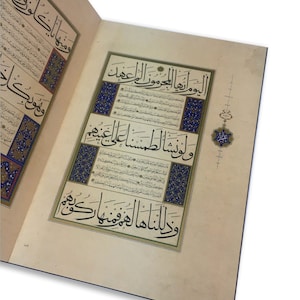 K&ouml;nnte beinhalten: Ein aufgeschlagenes Buch mit cremefarbenen Seiten, das eine komplexe schwarze arabische Kalligrafie zeigt. Der Text ist von dekorativen R&auml;ndern in Blau, Gold und Wei&szlig; umrahmt. Ein kleines, verziertes Blumenmotiv in Blau und Gold liegt am rechten Rand.