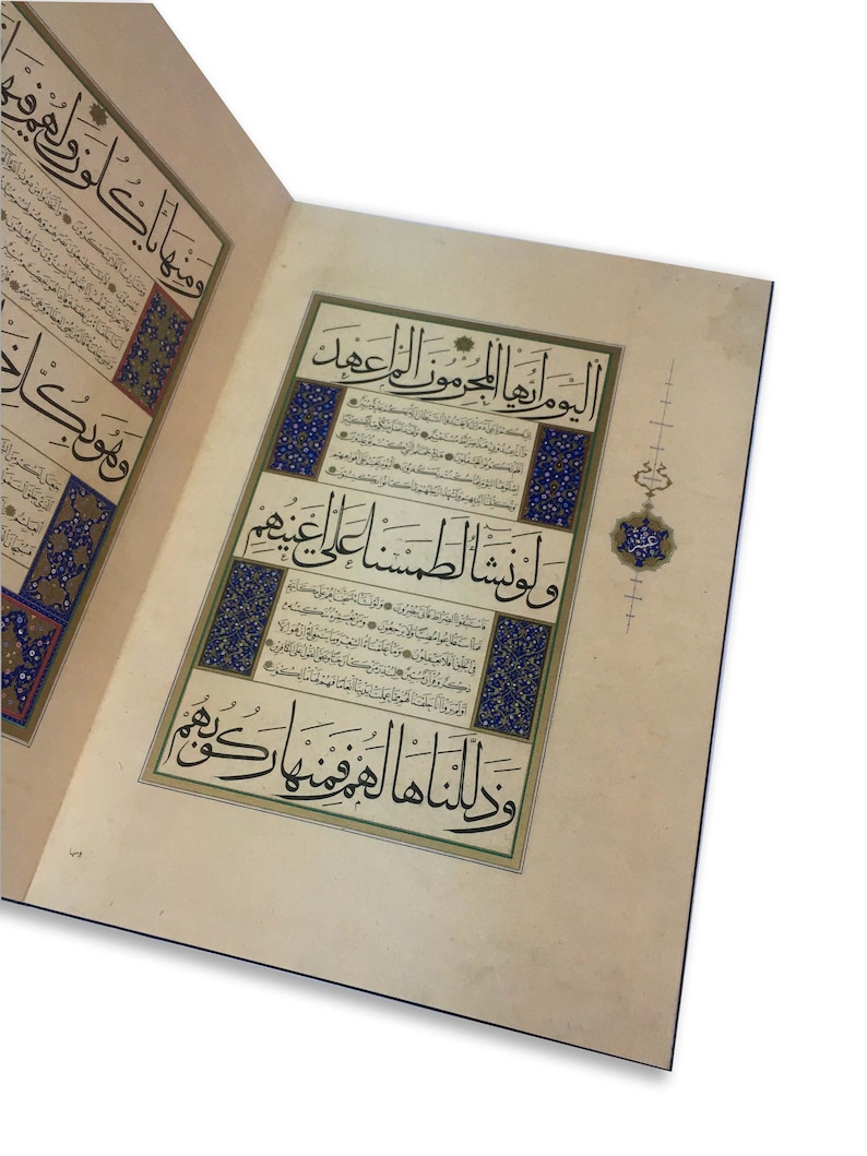 K&ouml;nnte beinhalten: Ein aufgeschlagenes Buch mit cremefarbenen Seiten, das eine komplexe schwarze arabische Kalligrafie zeigt. Der Text ist von dekorativen R&auml;ndern in Blau, Gold und Wei&szlig; umrahmt. Ein kleines, verziertes Blumenmotiv in Blau und Gold liegt am rechten Rand.