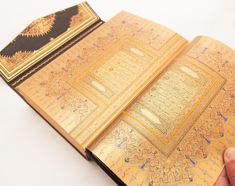 May include: An open book with a dark brown cover and gold-toned pages featuring intricate, ornate patterns and Arabic calligraphy. The pages are decorated with detailed gold filigree, geometric borders, and blue accents surrounding the central text.