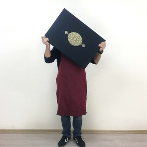 K&ouml;nnte beinhalten: Ein rechteckiges schwarzes Buch oder Etui mit einem zentralen goldfarbenen kreisf&ouml;rmigen Emblem mit komplexen Filigranmustern. Der Gegenstand wird horizontal gehalten und zeigt eine dunkle Oberfl&auml;che mit einem goldenen Design.