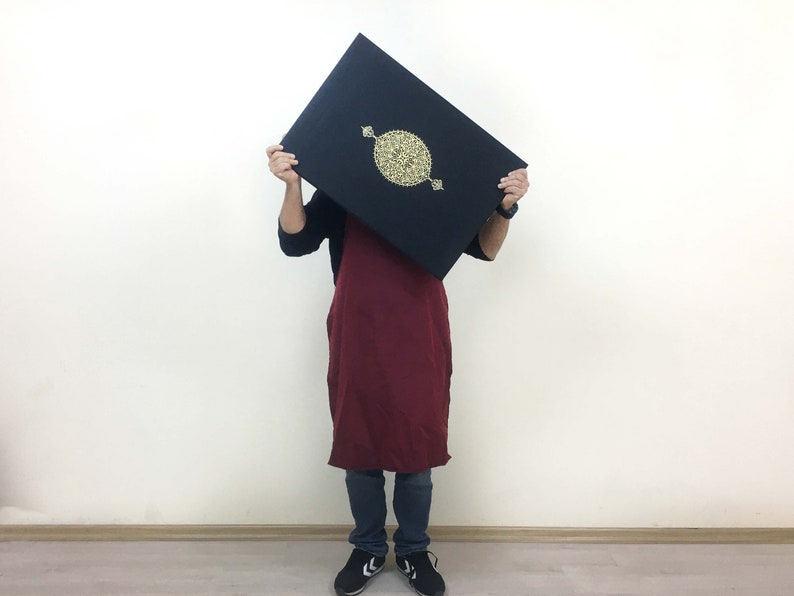 K&ouml;nnte beinhalten: Ein rechteckiges schwarzes Buch oder Etui mit einem zentralen goldfarbenen kreisf&ouml;rmigen Emblem mit komplexen Filigranmustern. Der Gegenstand wird horizontal gehalten und zeigt eine dunkle Oberfl&auml;che mit einem goldenen Design.