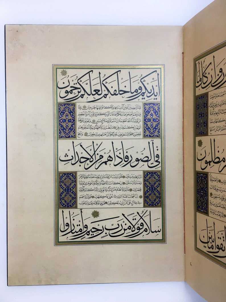 K&ouml;nnte beinhalten: Eine Seite aus einem illuminierten Manuskript mit komplexer arabischer Kalligrafie in schwarzer Tinte. Der Text ist von dekorativen R&auml;ndern mit blauen, goldenen und wei&szlig;en Blumenmustern umrahmt. Die Seite ist cremefarben und vertikal angelegt.