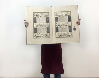 May include: A large, open book featuring two pages of intricate Arabic calligraphy in black ink. The text is framed by decorative blue and gold geometric borders. The pages are cream-colored with ornate floral motifs in the margins and center.