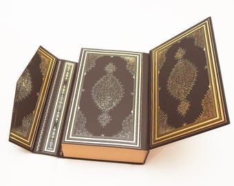 May include: A dark brown book with an open gatefold cover, featuring intricate gold foil filigree patterns on the interior panels and the front cover. The book has a rectangular shape with a thick spine and light-coloured page edges.