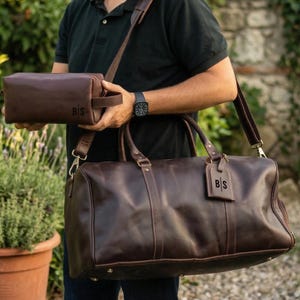 May include: A set of brown leather travel bags, including a duffel bag with a shoulder strap and a smaller toiletry bag. Both bags have the initials "B|S" embossed on them. The bags are a rich brown color and appear to be made of high-quality leather.