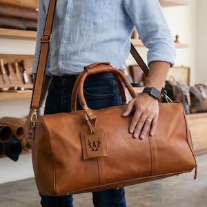 May include: A brown leather duffel bag with a shoulder strap and top handles. The bag has a luggage tag with a black emblem. The bag is held by a person wearing a light blue shirt and jeans.