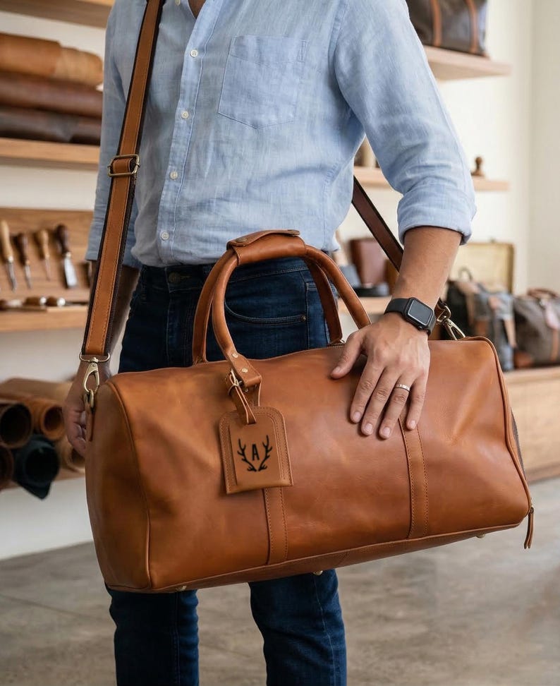 May include: A brown leather duffel bag with a shoulder strap and top handles. The bag has a luggage tag with a black emblem. The bag is held by a person wearing a light blue shirt and jeans.