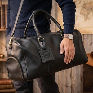 May include: Black leather duffel bag with a top handle and shoulder strap. The bag has a luggage tag with the initials "GH". The bag is held by a person. The bag is a travel accessory.