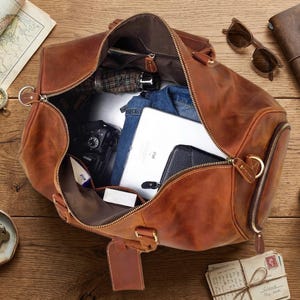 May include: A brown leather duffel bag, partially unzipped, revealing contents like a tablet, camera, and clothing. Accessories include sunglasses, a journal, a compass, and a cup of coffee. The items are arranged on a wooden surface.