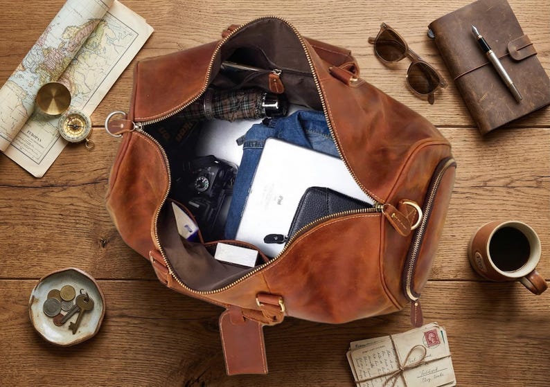 May include: A brown leather duffel bag, partially unzipped, revealing contents like a tablet, camera, and clothing. Accessories include sunglasses, a journal, a compass, and a cup of coffee. The items are arranged on a wooden surface.