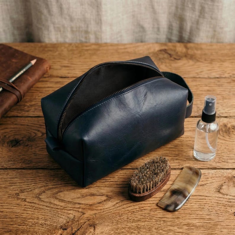 May include: A navy blue leather toiletry bag with a black zipper, open to reveal the interior. A small spray bottle, a wooden brush, and a comb are next to the bag. A leather-bound notebook and pencil are in the background.