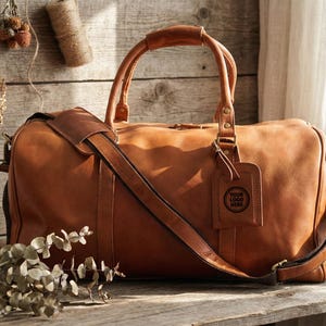 May include: A large, tan leather duffel bag with a shoulder strap and top handles. The bag has a luggage tag with a logo. The image also includes a small notebook, plants, and decorative items, all on a wooden surface.