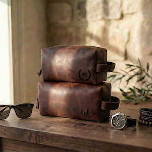 May include: Two brown leather toiletry bags with handles are stacked on a wooden surface. Sunglasses, a watch, and bracelets are also on the table. The bags have initials stamped on them. The background includes a window and a stone wall.