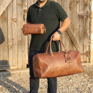 May include: A set of brown leather travel bags. The set includes a large duffel bag with handles and a smaller toiletry bag. Both bags have a circular logo tag. The bags are made of a smooth, rich brown leather.