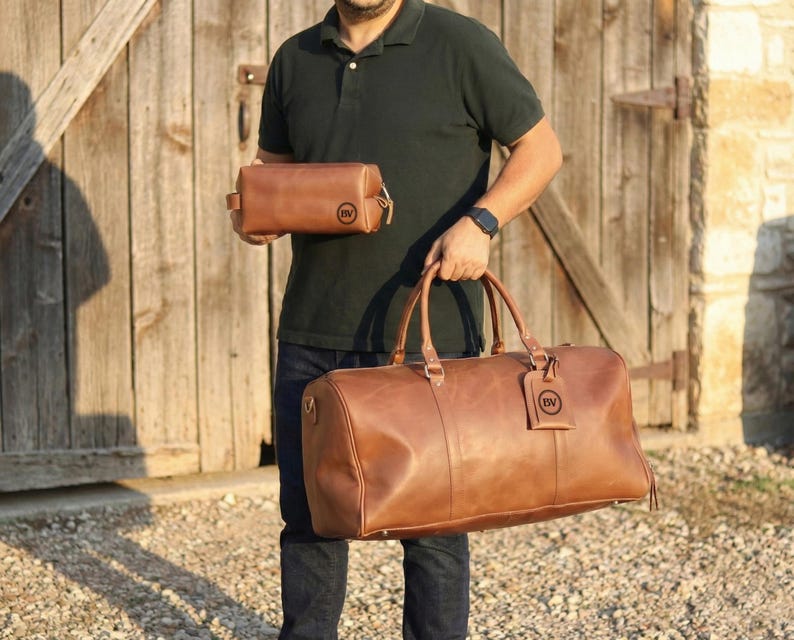 May include: A set of brown leather travel bags. The set includes a large duffel bag with handles and a smaller toiletry bag. Both bags have a circular logo tag. The bags are made of a smooth, rich brown leather.