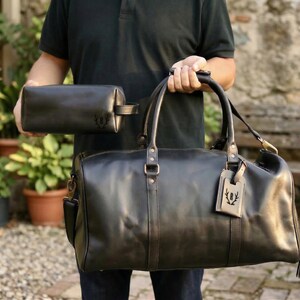 May include: A black leather duffel bag and matching toiletry bag are held up. The duffel bag has a shoulder strap and a luggage tag. Both bags have a debossed emblem. The bags are made of a smooth, dark leather.