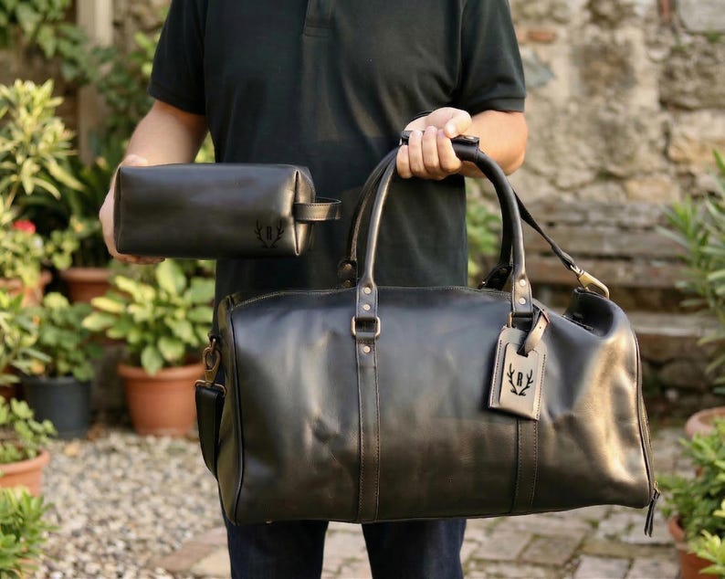 May include: A black leather duffel bag and matching toiletry bag are held up. The duffel bag has a shoulder strap and a luggage tag. Both bags have a debossed emblem. The bags are made of a smooth, dark leather.
