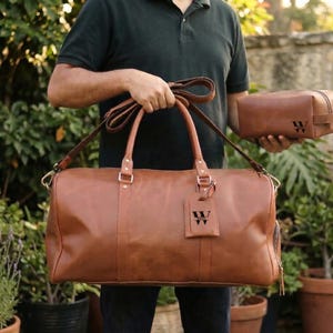 May include: A brown leather duffel bag with a matching toiletry bag. The duffel bag has a shoulder strap and carry handles. Both bags have the letter "W" embossed on them. The bags are held in front of a background of green foliage.