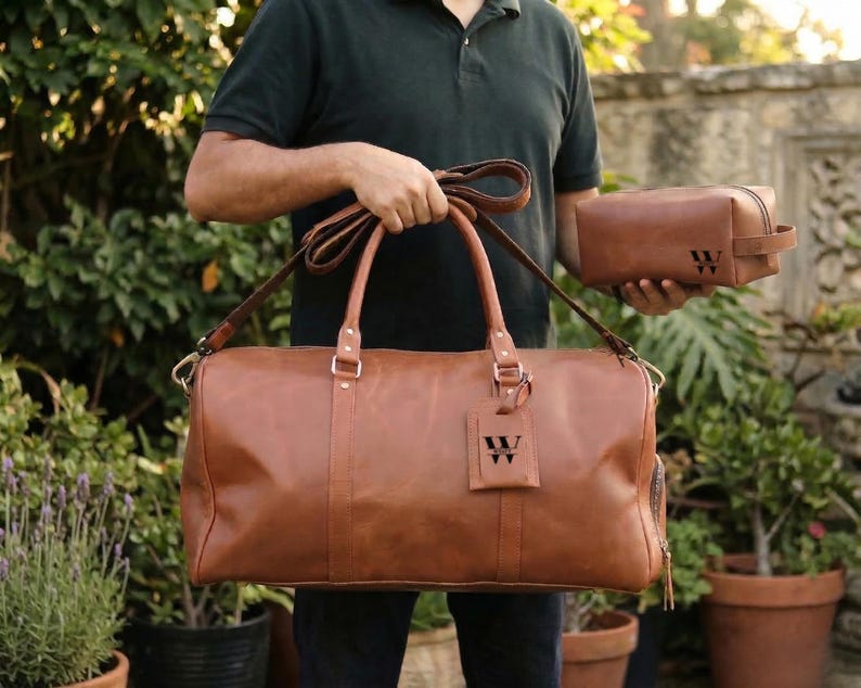 May include: A brown leather duffel bag with a matching toiletry bag. The duffel bag has a shoulder strap and carry handles. Both bags have the letter "W" embossed on them. The bags are held in front of a background of green foliage.