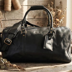 May include: A black leather duffel bag with a top handle and shoulder strap. The bag has a luggage tag that reads "LUKE". The bag is on a wooden surface.