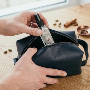 May include: A navy blue leather toiletry bag with a black zipper, holding a clear spray bottle labeled "Rebel Dark Spice." The bottle has a black spray nozzle and a decorative label. The bag is on a wooden surface.