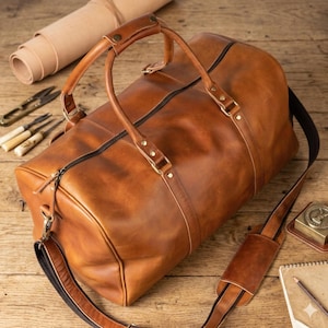 May include: A brown leather duffel bag with a black zipper and a detachable shoulder strap. The bag has two handles and metal hardware. The bag is on a wooden surface.