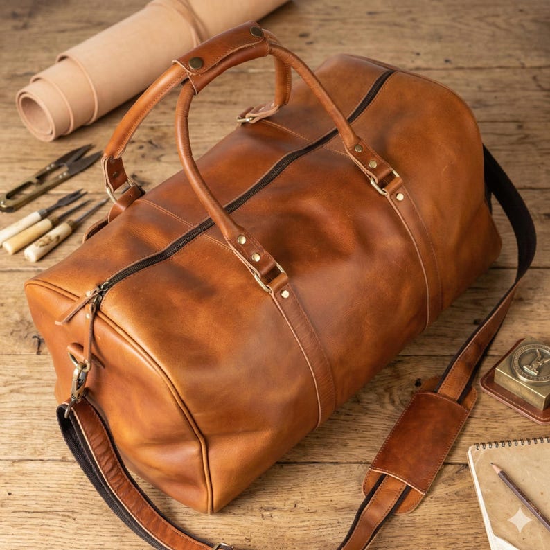May include: A brown leather duffel bag with a black zipper and a detachable shoulder strap. The bag has two handles and metal hardware. The bag is on a wooden surface.
