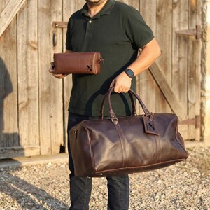 May include: A person holding a brown leather duffel bag and a matching toiletry bag. Both bags have a dark brown leather tag with the letter "N". The duffel bag has two handles and a shoulder strap.
