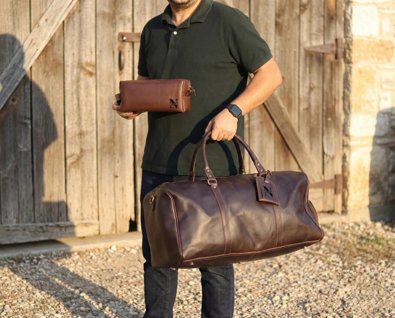May include: A person holding a brown leather duffel bag and a matching toiletry bag. Both bags have a dark brown leather tag with the letter "N". The duffel bag has two handles and a shoulder strap.