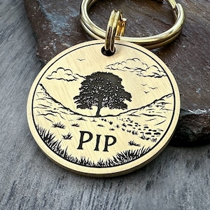 May include: A brass dog tag with a black engraved image of a tree in a valley with the word "PIP" underneath. The tag is attached to a gold key ring.