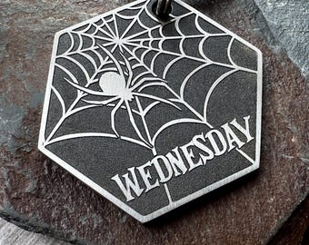 Könnte beinhalten: Ein sechseckiger Schlüsselanhänger aus Metall mit dunkelgrauer, texturierter Oberfläche. Das Design zeigt ein eingraviertes Spinnennetz, die Silhouette einer Spinne und das Wort WEDNESDAY in einer kräftigen, stilisierten Schriftart.