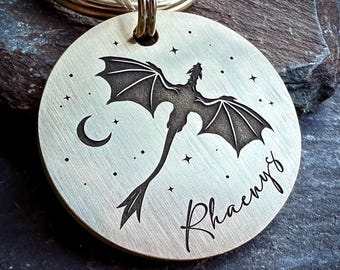 May include: A circular brass-toned metal keychain tag featuring a laser-engraved silhouette of a dragon with outstretched wings, a crescent moon, and scattered stars. The name Rhaenys is engraved in a cursive script font on the lower right side.
