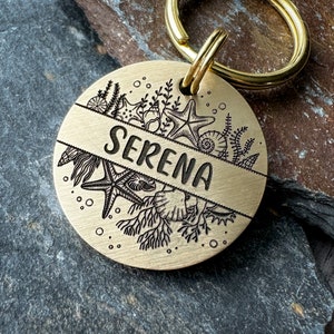 Op de afbeelding: Een messing hondenpenning met een zeeleven-ontwerp en de naam "SERENA" erop gegraveerd.