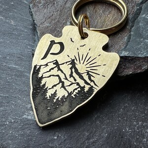 Può includere: Un portachiavi a forma di freccia in ottone con un design di montagna e foresta. Il design include la lettera "P", una catena montuosa, un sole e alberi. Il portachiavi ha un anello color oro.