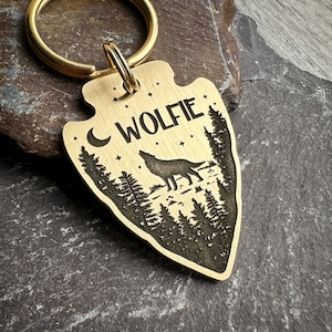 Op de afbeelding: Een gouden pijlpuntvormige sleutelhanger met een wolf die huilt naar de maan in een bos scène. De sleutelhanger heeft de naam "WOLFIE" erop gegraveerd.