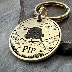 May include: Brass dog tag with a laser engraved design of a tree, hills, and paw prints. The tag reads "PIP".
