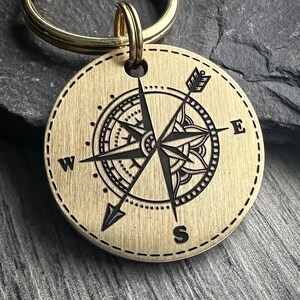 Op de afbeelding: Een messing kompas sleutelhanger met een gedetailleerd kompas ontwerp en de letters W, E, S en N gegraveerd op het kompas. De sleutelhanger heeft een gouden metalen ring.