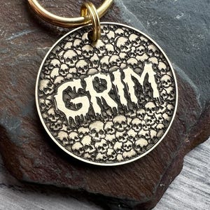 May include: A round, brass-colored pet tag with the word "GRIM" in a dripping font, surrounded by a pattern of small skulls. The tag is attached to a gold-colored ring.