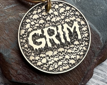 May include: A circular, brass-toned pet tag featuring the word "GRIM" in a dripping font, set against a backdrop of tiny skulls. The tag is connected to a gold-coloured ring.