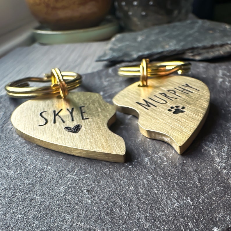 Puede incluir: Dos medallas para perros en forma de coraz&oacute;n de oro con anillos partidos. La medalla de la izquierda dice "Skye" con un peque&ntilde;o s&iacute;mbolo de coraz&oacute;n. La medalla de la derecha dice "Murphy" con un s&iacute;mbolo de huella de pata.