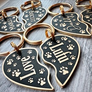 May include: A set of five gold-coloured heart-shaped dog tags with black enamel and gold lettering. The tags feature the names "Marley", "Biscuit", "Paddy", "Love", and "Luffu". Each tag has a gold ring for attaching to a collar or leash.