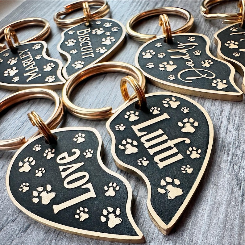May include: A set of five gold-coloured heart-shaped dog tags with black enamel and gold lettering. The tags feature the names "Marley", "Biscuit", "Paddy", "Love", and "Luffu". Each tag has a gold ring for attaching to a collar or leash.