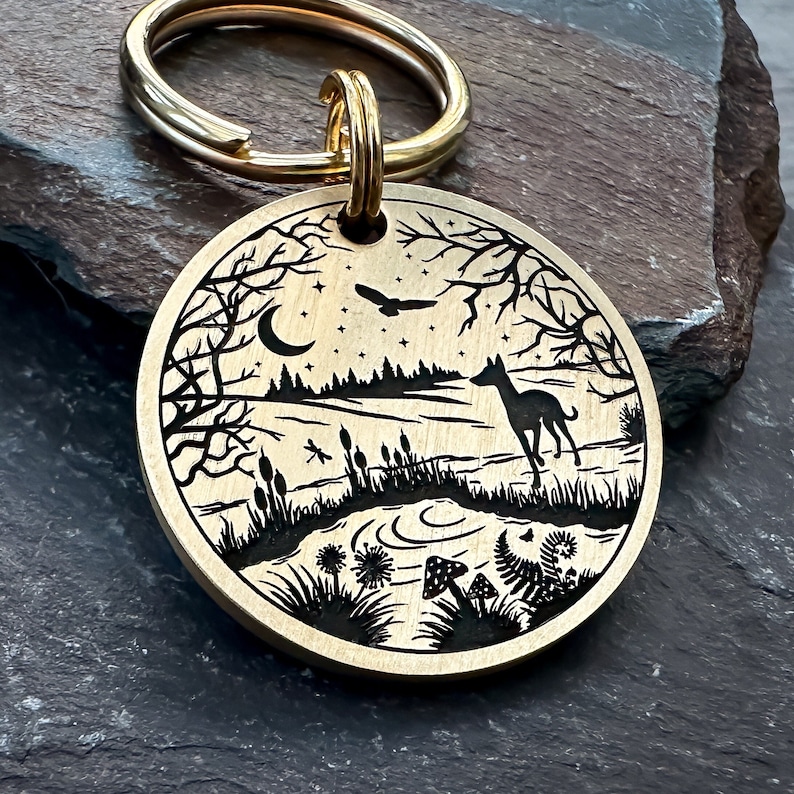 May include: A gold-toned metal key chain with a round tag featuring a black and white engraved design of a forest scene with a deer, a dragonfly, a crescent moon, stars, and mushrooms.
