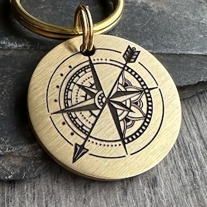 Op de afbeelding: Een gouden kompas sleutelhanger met een zwart kompas ontwerp en een pijl die naar het noorden wijst. Het kompas is omgeven door een decoratieve rand.