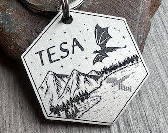 May include: A hexagonal silver-toned metal keychain tag featuring an engraved landscape scene with a flying dragon, a mountain range, a forest, and a lake. The name TESA is engraved in black capital letters, surrounded by small star symbols.