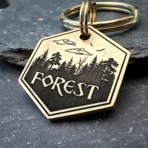 May include: A brass key chain with a hexagonal tag featuring a silhouette of a forest with trees, clouds, and birds. The tag is engraved with the word "FOREST".
