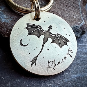 Könnte beinhalten: Ein silberfarbener Hundemarkenanhänger aus Metall mit einer schwarzen Silhouette eines Drachen, der vor einem Halbmond und Sternen fliegt. Der Name "Rhaenyra" ist unter dem Drachen eingraviert.