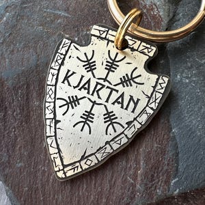 May include: A silver arrowhead-shaped keyring with the name "KJARTAN" in black lettering. The design features a central symbol and border runes. A gold-coloured ring is attached for keys.
